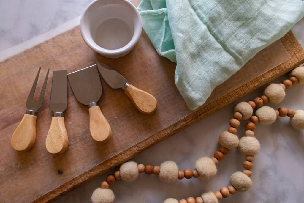 How to Assemble a Charcuterie Board by popular Utah craft blog, Sweet Red Poppy: image of a wooden board, cheese knives with wooden handles, teal tea towel, wood bead and felt pom garland, and white ceramic bowl. 