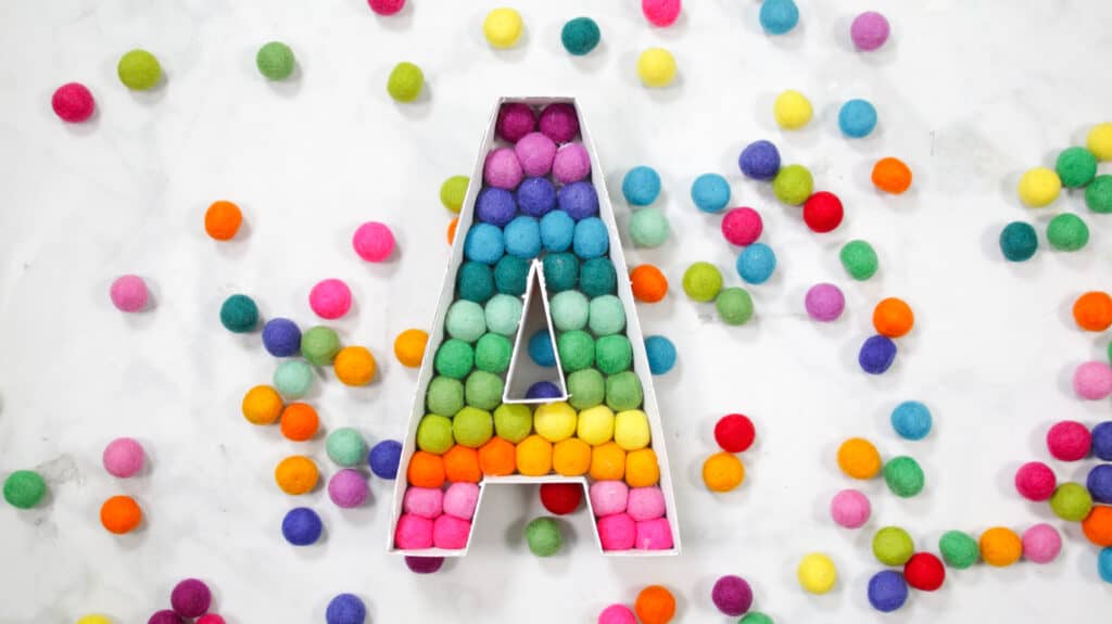 Felt Ball Letters by popular US craft blog, Sweet Red Poppy: image of a white cardboard letter A filled with rainbow felt balls. 