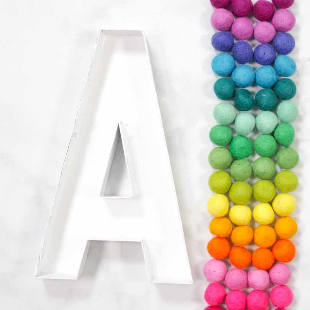 Felt Ball Letters by popular US craft blog, Sweet Red Poppy: image of a white cardboard letter A next to a row  of rainbow felt balls. 