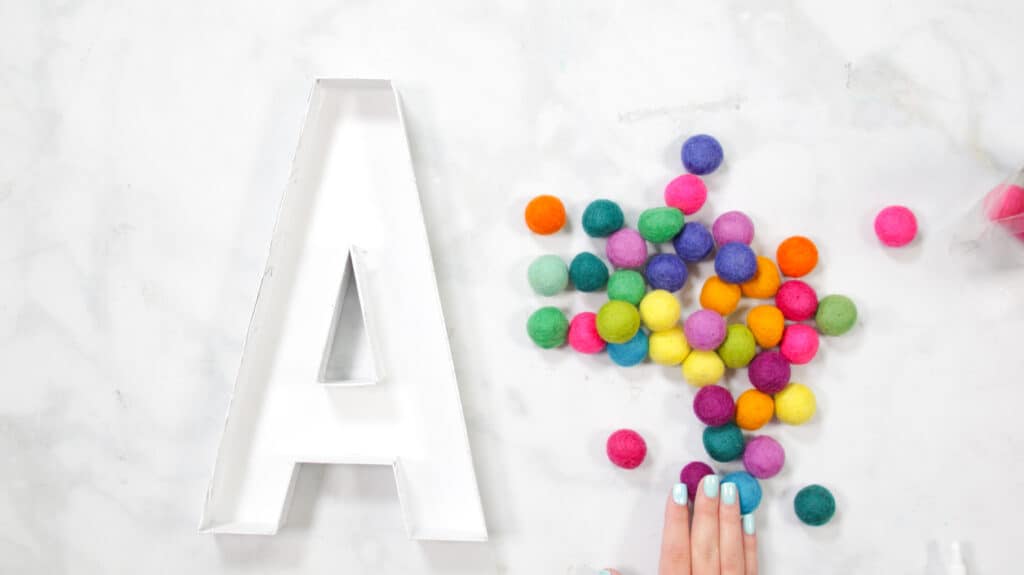 Felt Ball Letters by popular US craft blog, Sweet Red Poppy: image of a white cardboard letter A next to a pile of rainbow felt balls. 