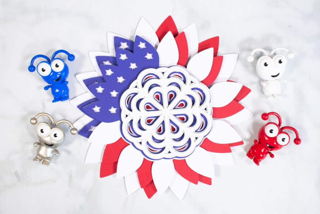 DIY 4th of July Wreath by popular US craft blog, Sweet Red Poppy: image of a layered DIY 4th of July wreath surrounded by some red, white, blue, and silver Cricut mascots. 