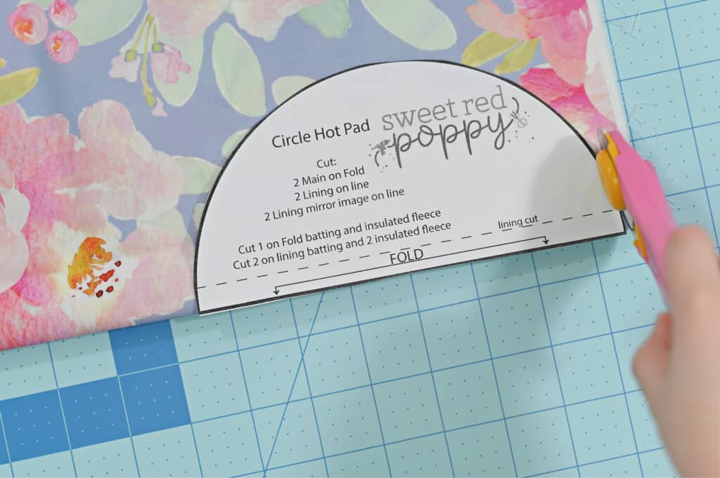 Oven Mitt Sewing Pattern by popular US sewing blog, Sweet Red Poppy: image of a circle hot pad pattern resting on top of some floral fabric and being cut out with a pink rotary cutter on a blue rotary cutting board. 