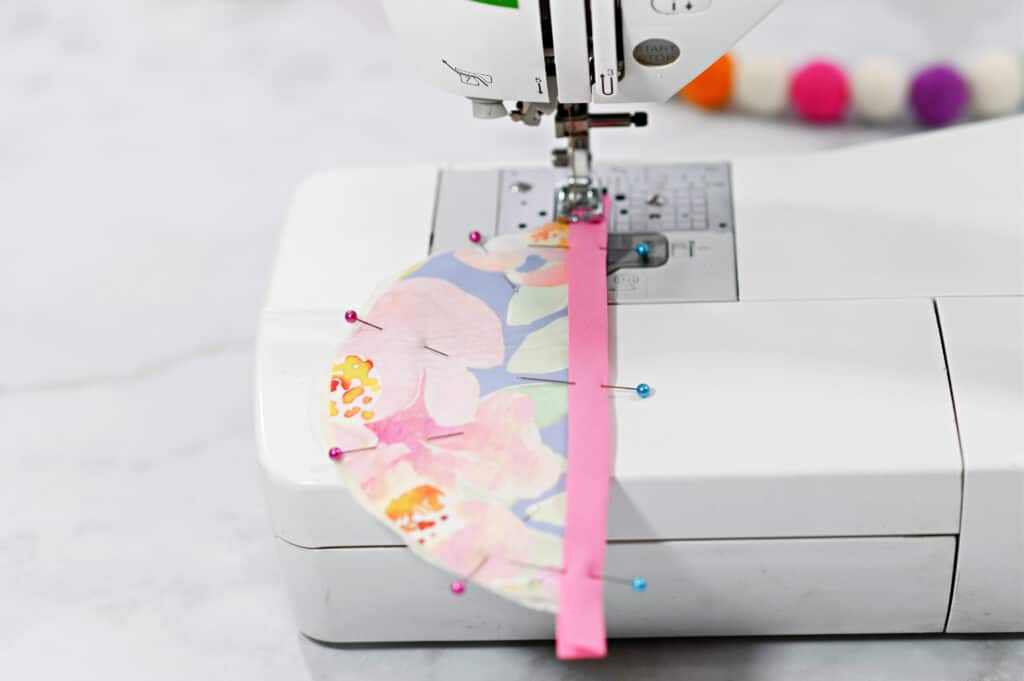 Oven Mitt Sewing Pattern by popular US sewing blog, Sweet Red Poppy: image of a floral print oven mitt pinned together with fabric pins being sewn on a sewing machine. 