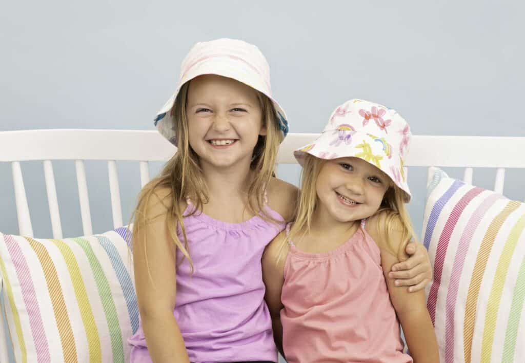 Bucket Hat Sewing Pattern by popular US sewing blog, Sweet Red Poppy: image of two girls sitting on a white wooden bench with rainbow stripe throw pillows and wearing bucket hats. 