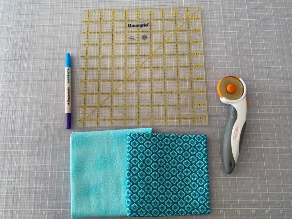 two fabrics, a ruler, a rotary cutter, and a fabric marking pen on a grey cutting mat
